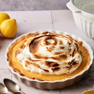 A Lemon Meringue Pie in a white pie dish sits on a tiled surface, surrounded by two lemons, a mixing bowl, three spoons, and a lemon squeezer.
