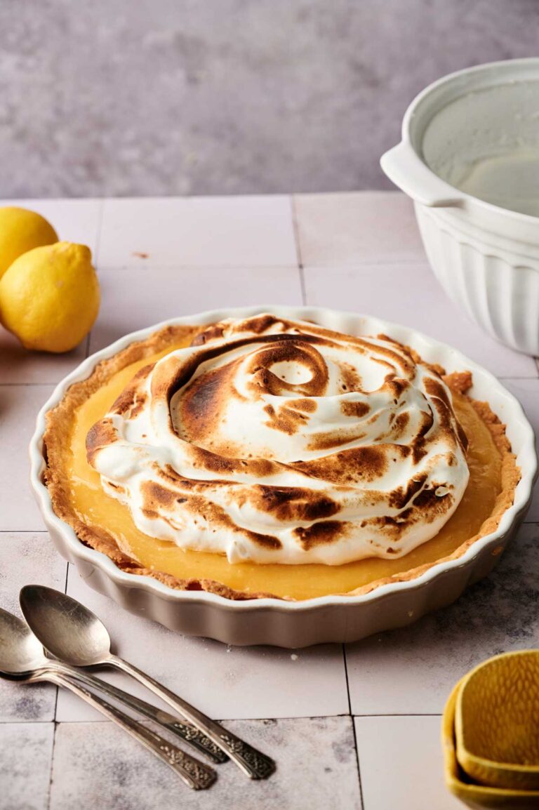 A Lemon Meringue Pie in a white pie dish sits on a tiled surface, surrounded by two lemons, a mixing bowl, three spoons, and a lemon squeezer.