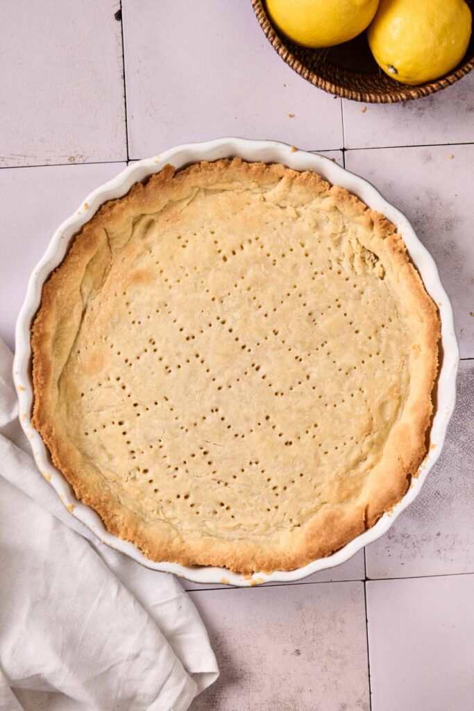 A baked pie crust with fork marks in a white dish sits on a tiled surface next to a basket of lemons, awaiting its transformation into a classic Lemon Meringue Pie. A white cloth adds a touch of elegance to the scene.