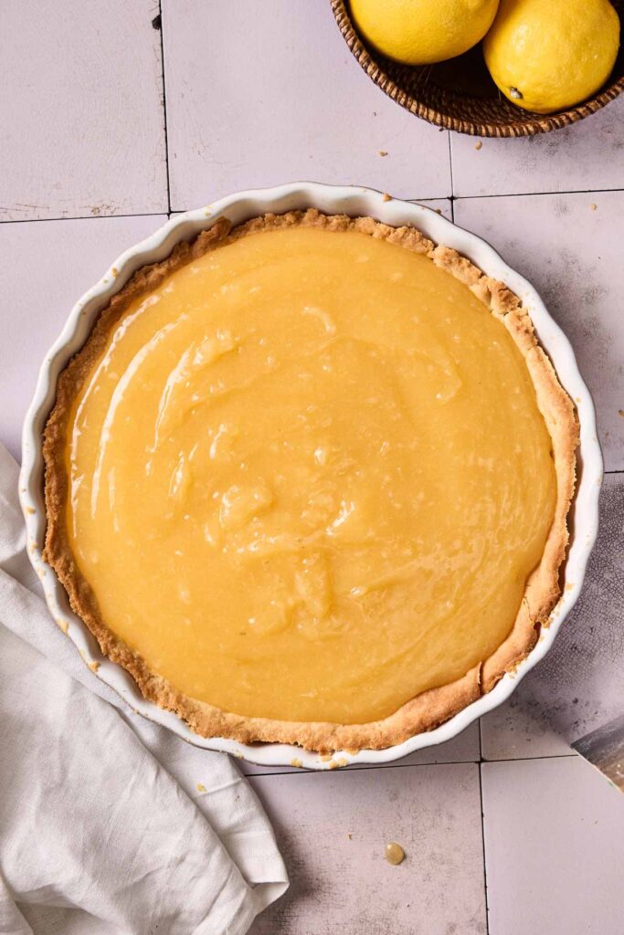 A baked pie crust filled with smooth lemon curd sits on a tiled surface next to a white cloth and a bowl of lemons, ready to become the perfect Lemon Meringue Pie.