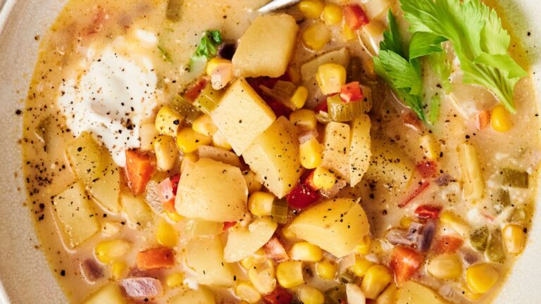 A close-up of a vegetable chowder with potatoes, corn, diced vegetables, a dollop of cream, black pepper, and a sprig of celery on top.