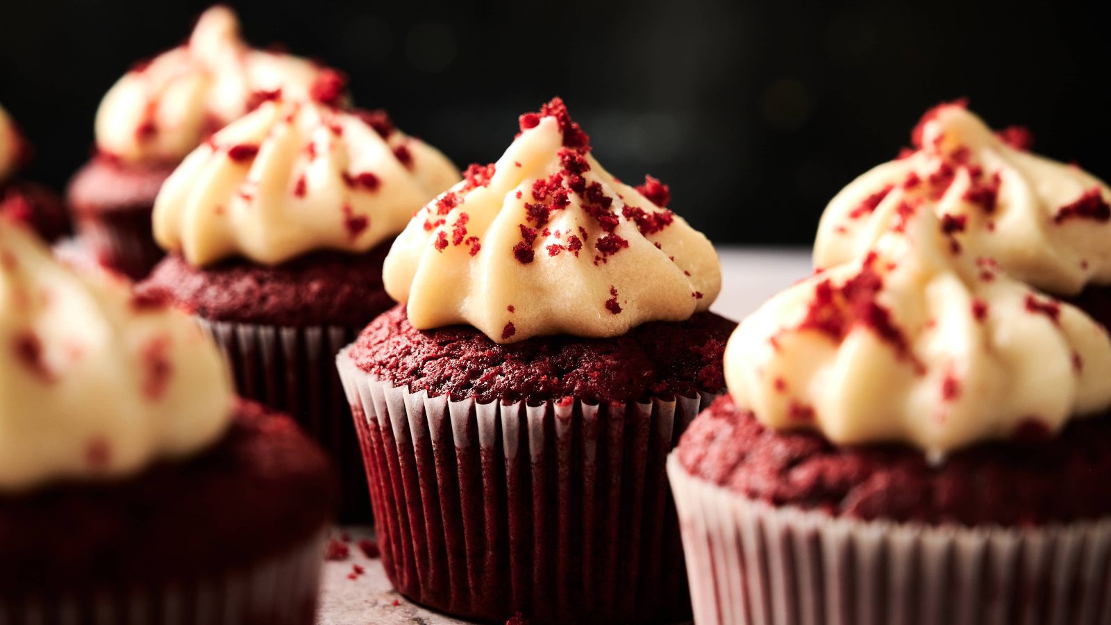 Close-up of several red velvet cupcakes topped with cream cheese frosting and red cake crumbs, arranged in rows on a surface.