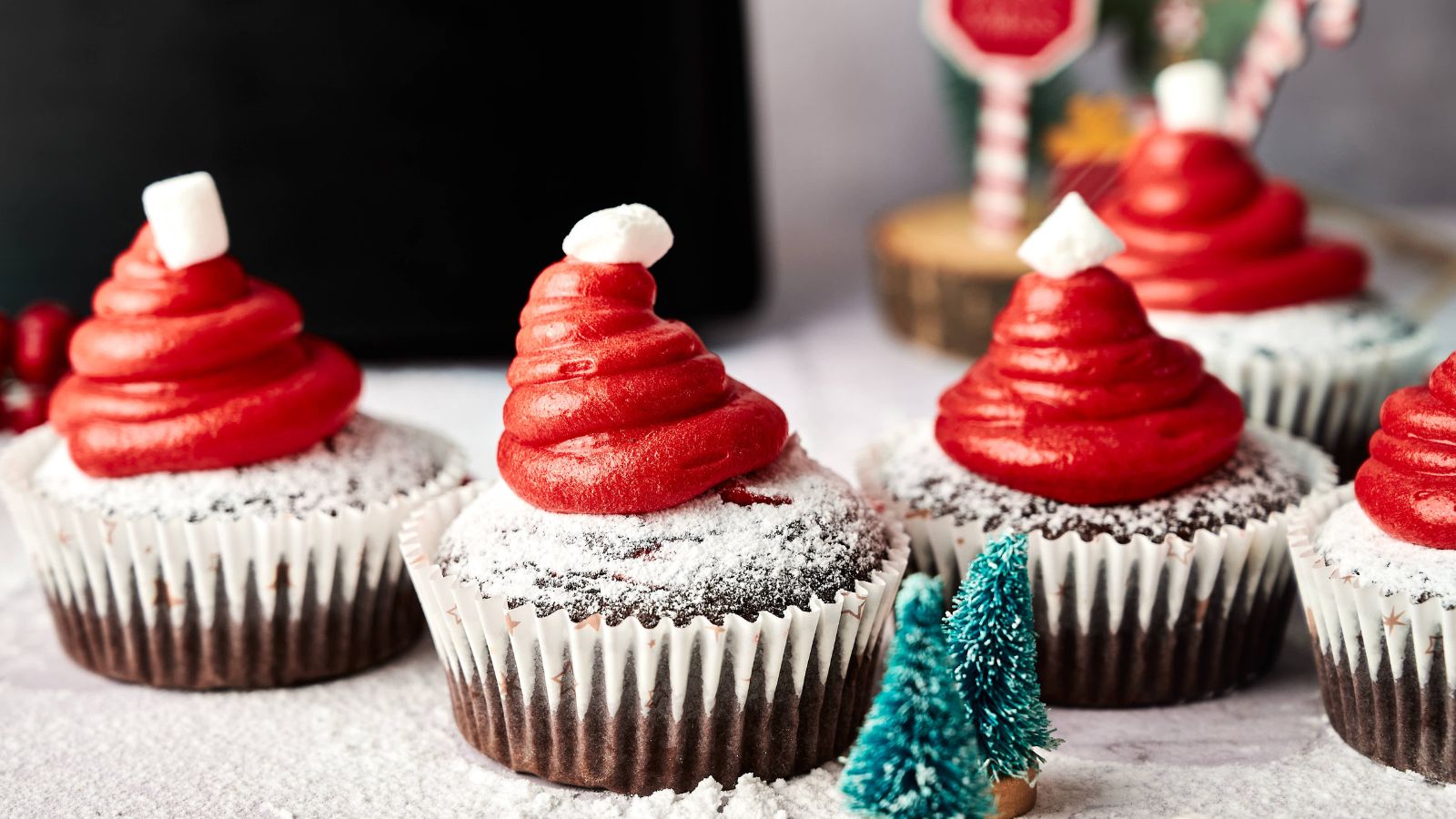 Chocolate cupcakes topped with red frosting shaped like Santa hats, each with a small marshmallow on top, dusted with powdered sugar, and a miniature Christmas tree in front.