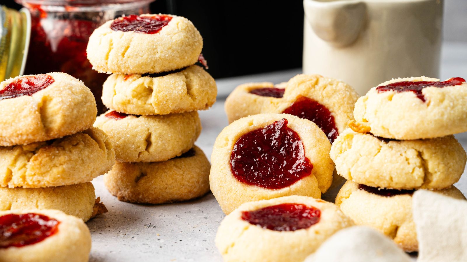 Stacks of thumbprint cookies filled with red jam are arranged on a light countertop, with a jar of jam and a cream-colored pitcher in the background.