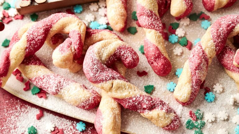 Candy cane-shaped cookies with red and white twisted dough, dusted with powdered sugar, are arranged on a board with festive sprinkles shaped like snowflakes and holly.