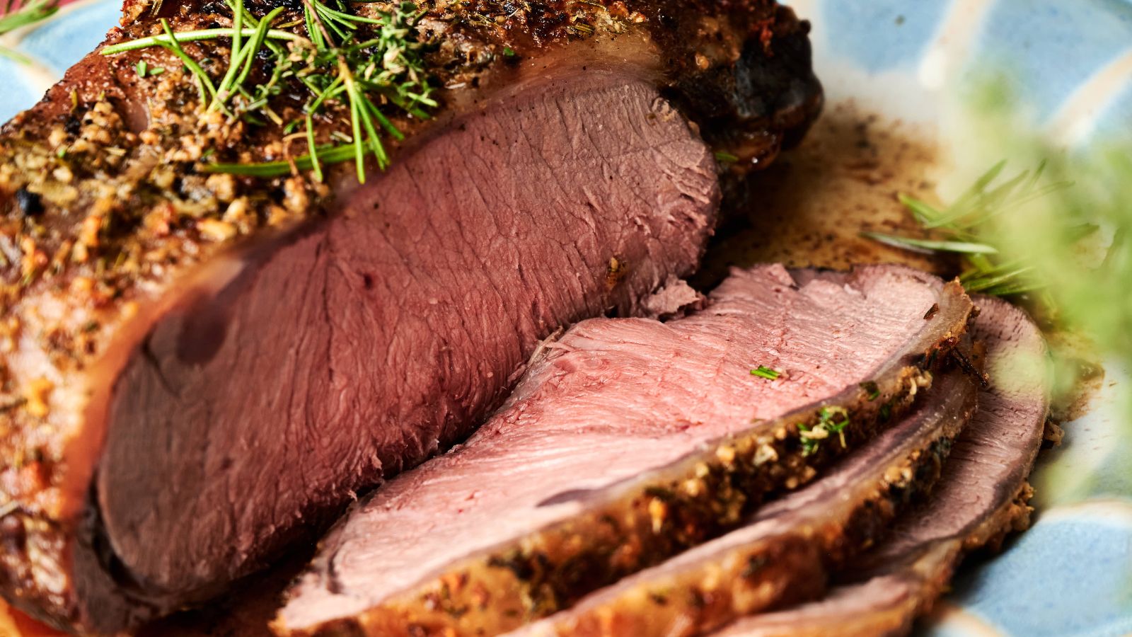 Close-up of a sliced roast beef garnished with herbs on a blue plate. The meat appears medium-rare and juicy.