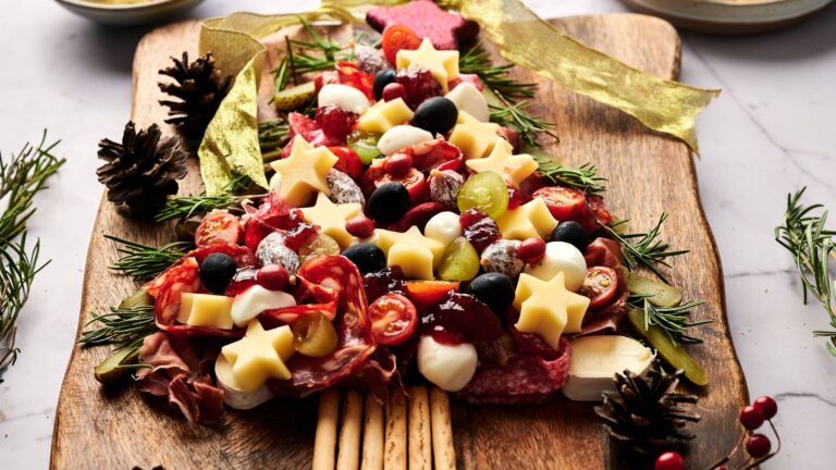 A Christmas tree-shaped charcuterie board with meats, cheeses, olives, vegetables, and herbs on a wooden tray, decorated with festive garnishes.