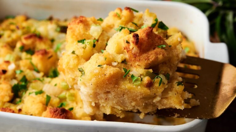 A close-up of a serving of baked stuffing with chopped parsley, scooped from a white baking dish with a metal spatula.