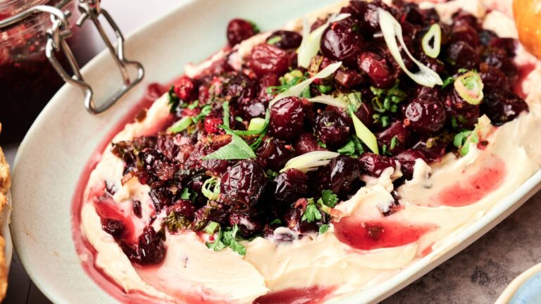 A platter of creamy cheese spread topped with roasted cranberries, chopped herbs, and sliced green onions.