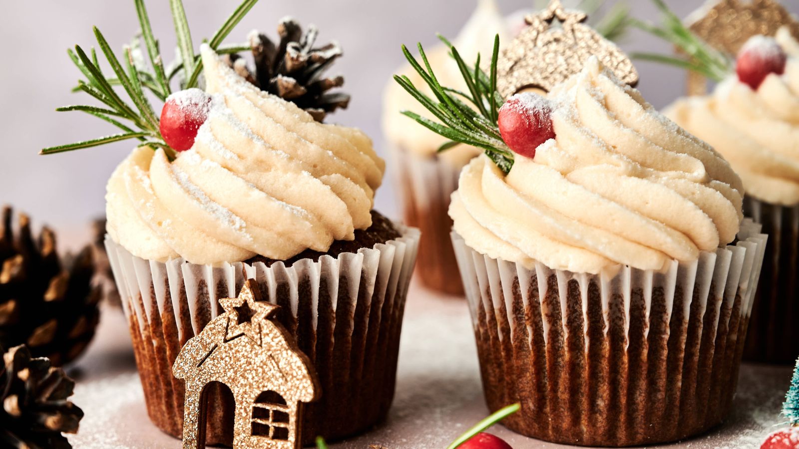 Three cupcakes topped with swirled frosting, cranberries, rosemary sprigs, pinecones, and powdered sugar, with a small decorative house in the foreground.