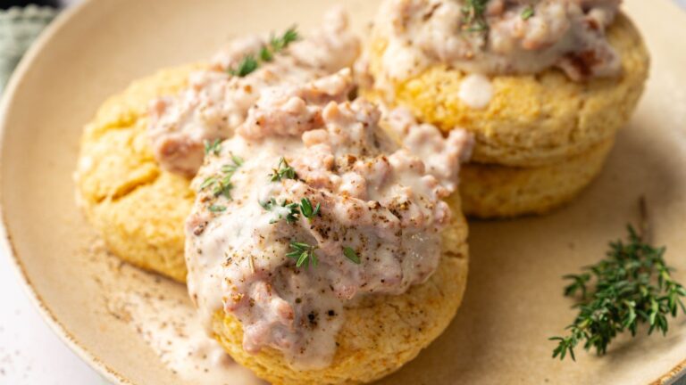 Two biscuits topped with sausage gravy and garnished with fresh thyme are served on a beige plate.