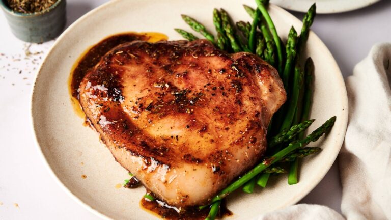 A cooked pork chop with sauce is served on a plate alongside steamed asparagus spears.