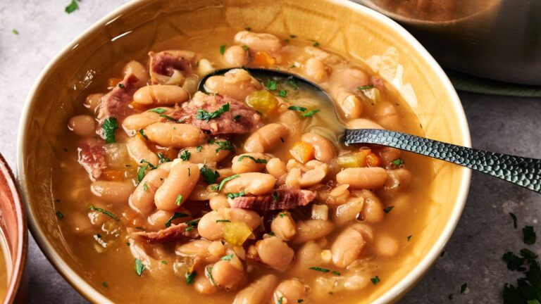 A bowl of bean soup with chunks of ham, garnished with chopped parsley, with a spoon resting in the bowl.