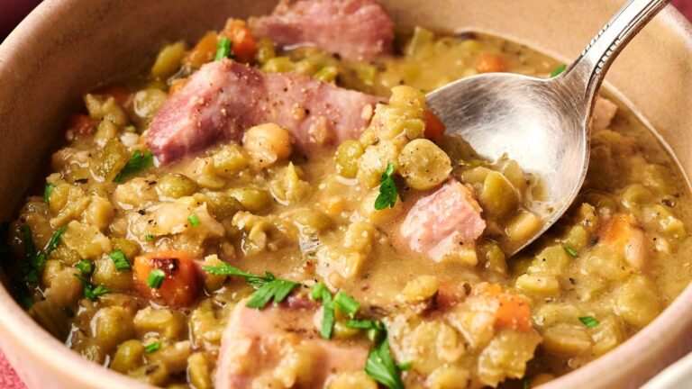 A bowl of split pea soup with chunks of ham, diced carrots, and green herbs, with a spoon resting in the soup.