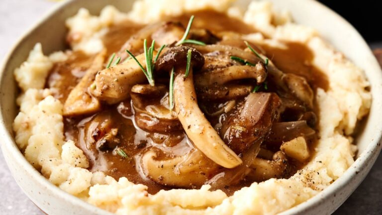 A bowl of mashed potatoes topped with mushroom gravy, garnished with fresh rosemary and ground black pepper.