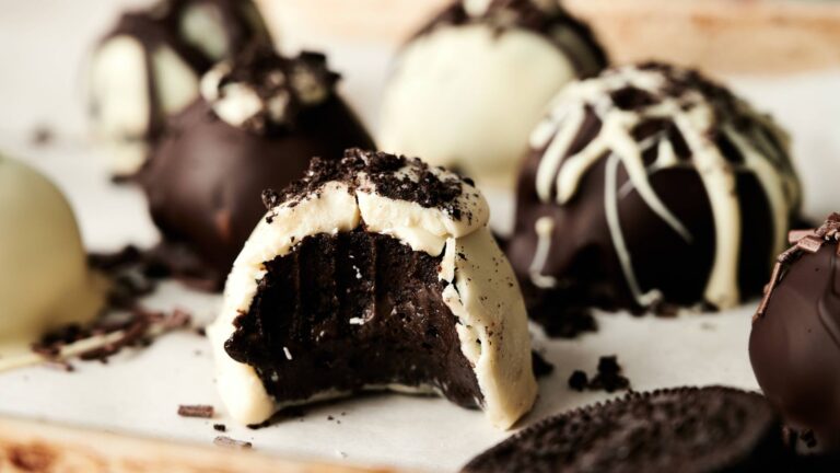 Close-up of assorted chocolate truffles, one with a bite taken out revealing a dark chocolate filling, topped with cookie crumbs and white chocolate drizzle.