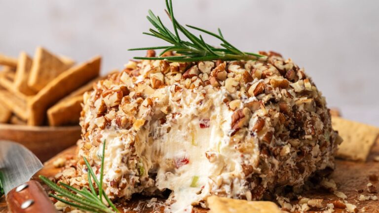 A cheese ball coated in chopped nuts, garnished with a sprig of rosemary, with crackers and a serving knife on the side.