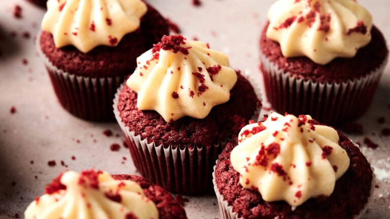 Several red velvet cupcakes topped with swirls of cream cheese frosting and sprinkled with red crumbs are arranged on a light surface.