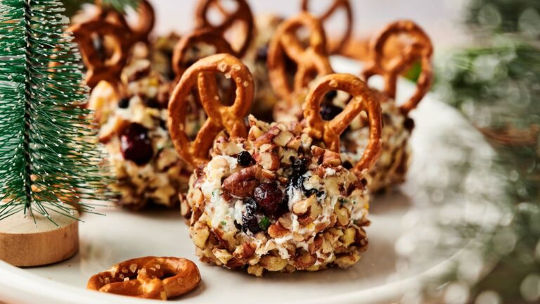 A plate of cheese balls coated in chopped nuts, garnished with dried fruit and pretzels, presented on a white surface with artificial pine tree decorations.