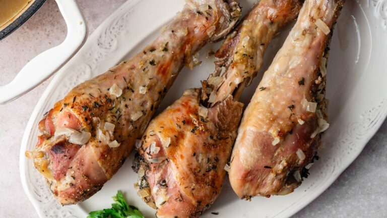 Three roasted turkey legs seasoned with herbs and diced onions are arranged on a white platter, with a small bunch of parsley on the side.