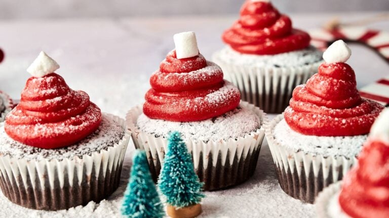 Chocolate cupcakes topped with red frosting, powdered sugar, and a small marshmallow, arranged to resemble Santa hats, with decorative miniature trees in the foreground.