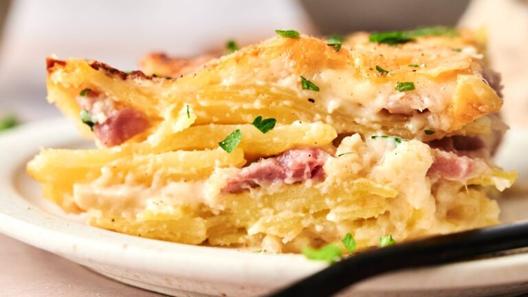 A close-up of a serving of potato gratin with ham, cheese, and creamy sauce, garnished with chopped parsley on a white plate.