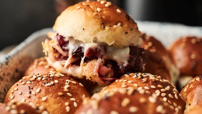 A close-up of a tray of poppy and sesame seed slider buns, with one sandwich filled with shredded meat, melted cheese, and cranberry sauce.