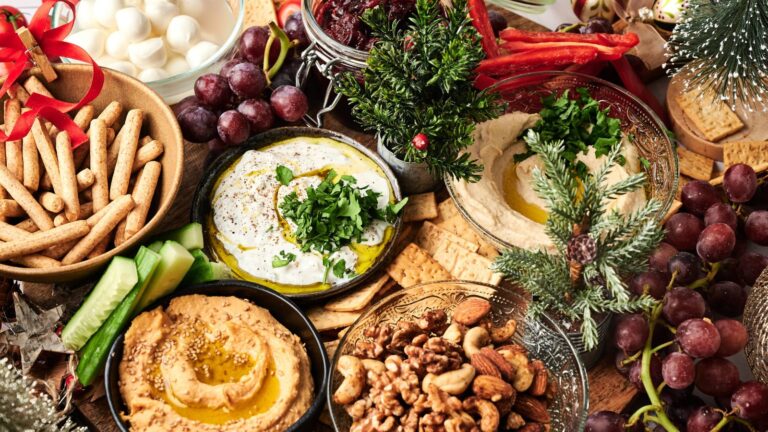 A holiday-themed spread with hummus, yogurt dip, breadsticks, nuts, crackers, grapes, mozzarella balls, and festive greenery decorations.