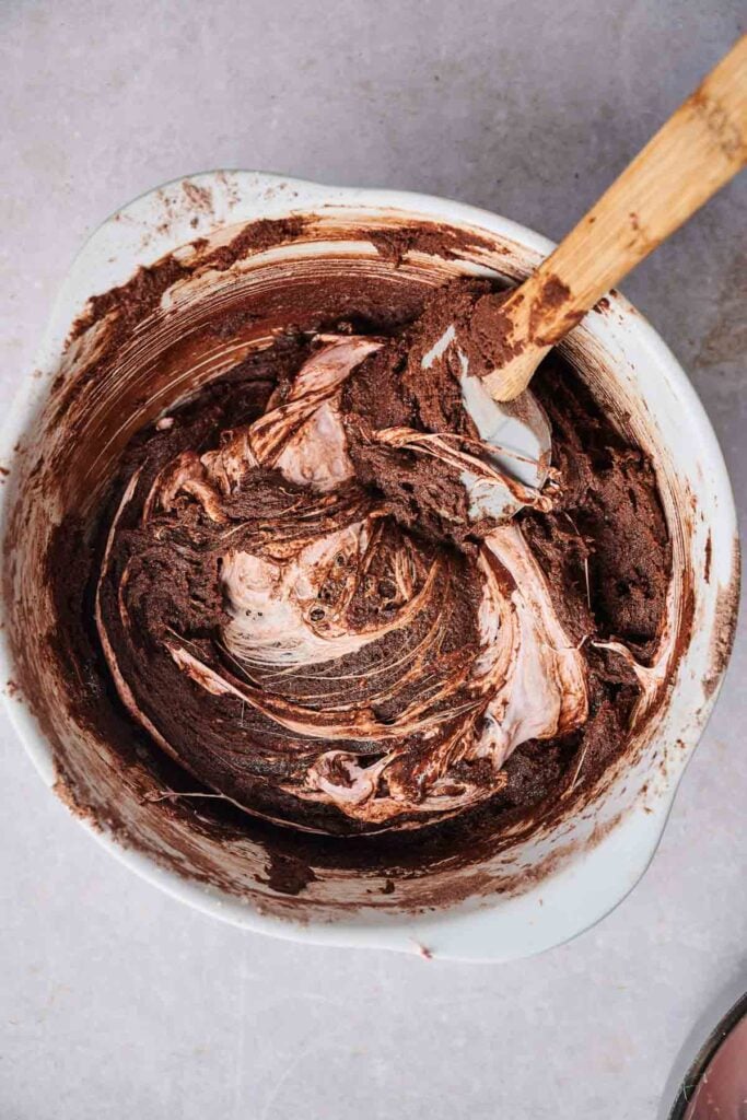 A mixing bowl containing chocolate batter being folded with a spatula, with visible streaks of lighter cream or meringue&mdash;perfect for creating Air Fryer Marshmallow Swirl Cookies.