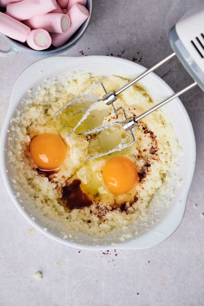 A mixing bowl with creamed butter and sugar, two cracked eggs, and vanilla extract is being mixed with an electric hand mixer&mdash;perfect for making Air Fryer Marshmallow Swirl Cookies. A bowl of pink marshmallows sits nearby, ready to swirl in.