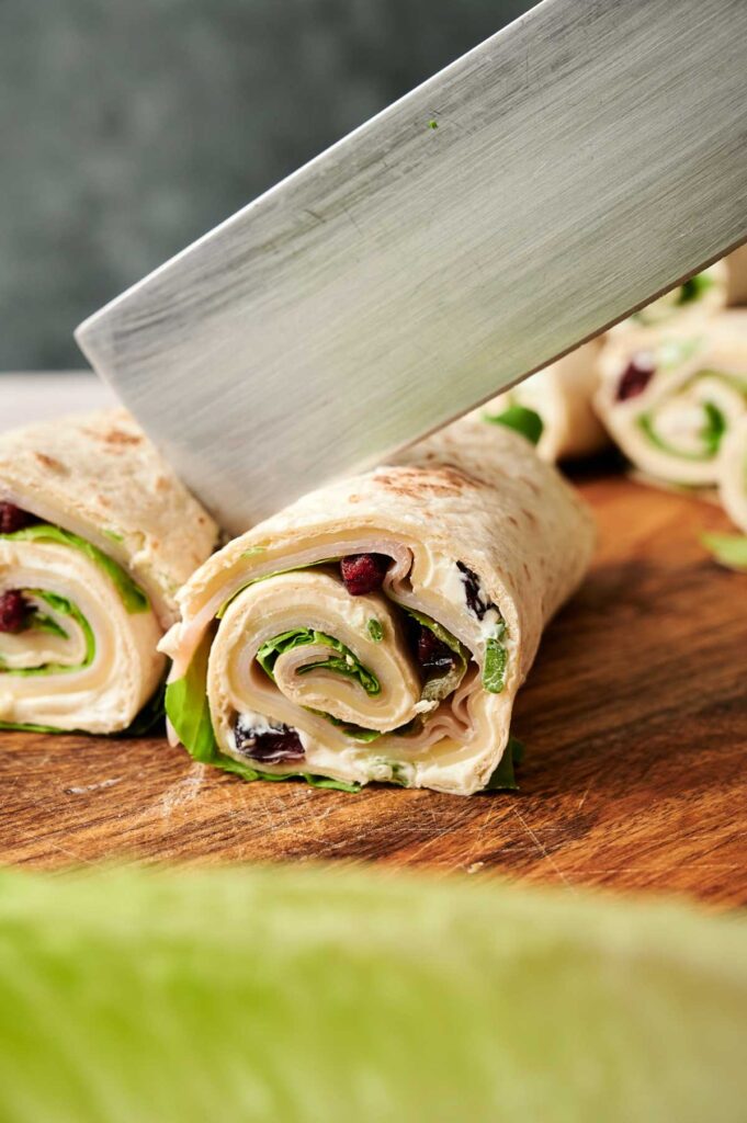 A large knife slices a rolled Turkey Pinwheels tortilla wrap filled with turkey, lettuce, and cranberry on a wooden cutting board.