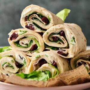 A plate with several Turkey Pinwheels—rolled tortilla wraps filled with lettuce, sliced cheese, and dried cranberries—stacked on top of each other.