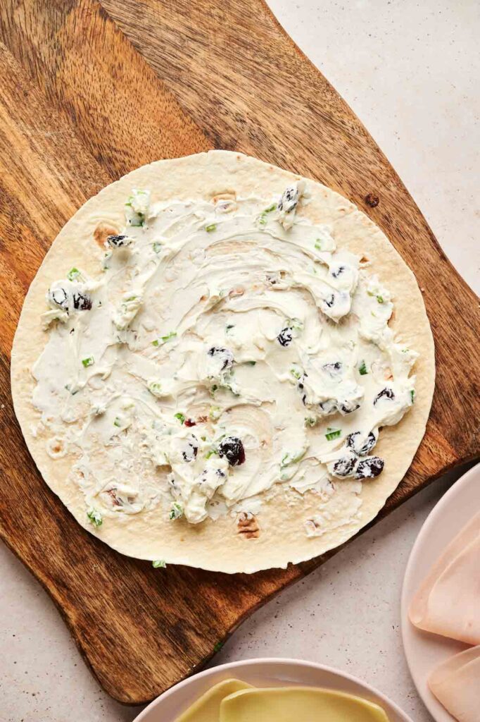 A flour tortilla on a wooden board is spread with a cream cheese mixture containing chopped green onions and dried cranberries—perfect for assembling delicious Turkey Pinwheels.