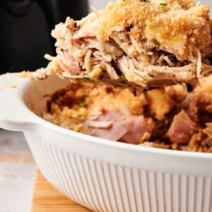 A serving of shredded, baked chicken with a breadcrumb topping is being lifted from a white casserole dish on a wooden surface—perfect for fans of Air Fryer Chicken Cordon Bleu Casserole.