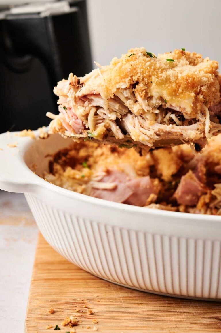 A serving of shredded, baked chicken with a breadcrumb topping is being lifted from a white casserole dish on a wooden surface—perfect for fans of Air Fryer Chicken Cordon Bleu Casserole.