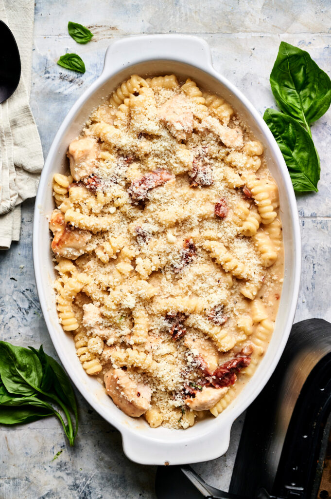 Oval white dish filled with creamy Air Fryer Marry Me Chicken Casserole, featuring pasta, pieces of chicken, sun-dried tomatoes, and grated cheese. Fresh basil leaves are placed nearby on the table.