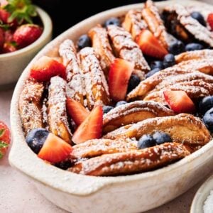 A ceramic baking dish filled with baked French toast slices, topped with powdered sugar, strawberries, and blueberries&mdash;perfect for serving alongside an Air Fryer Pancake Casserole at brunch.