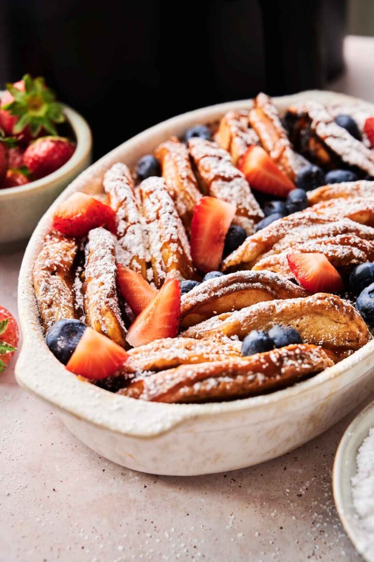 A ceramic baking dish filled with baked French toast slices, topped with powdered sugar, strawberries, and blueberries—perfect for serving alongside an Air Fryer Pancake Casserole at brunch.