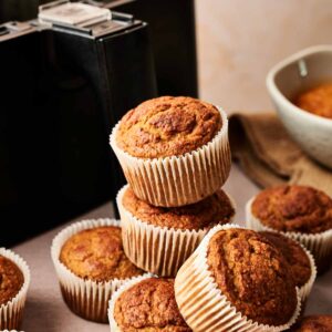 A stack of golden-brown Air Fryer Pumpkin Muffins in paper liners sits in front of an air fryer and a bowl on a beige surface.