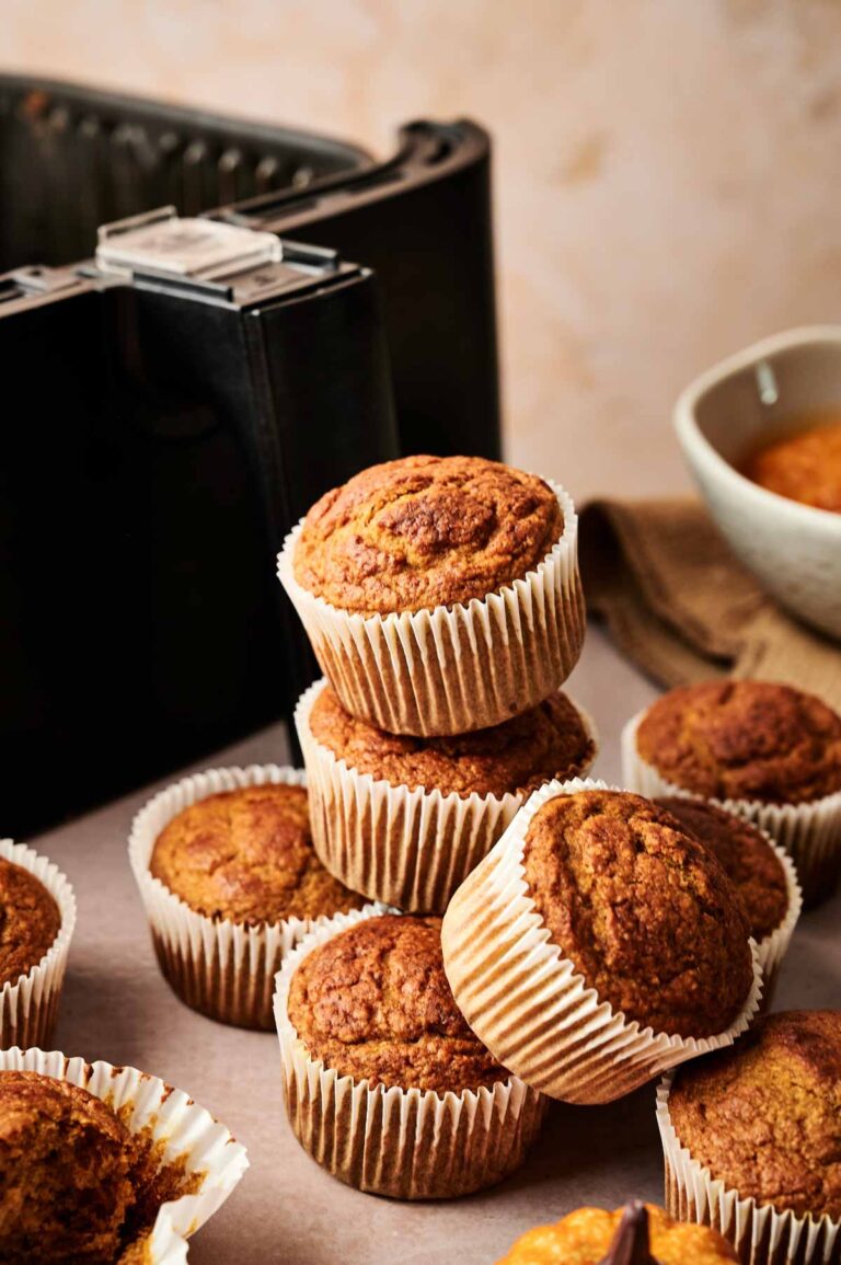 A stack of golden-brown Air Fryer Pumpkin Muffins in paper liners sits in front of an air fryer and a bowl on a beige surface.