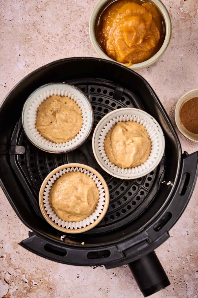 Three muffin liners filled with Air Fryer Pumpkin Muffins batter sit in an air fryer basket. A bowl of pumpkin puree and a small bowl of cinnamon are nearby on a light-colored surface.