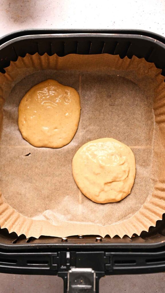 Two round portions of Air Fryer Pumpkin Pancake batter on parchment paper inside an air fryer basket, ready to be cooked.