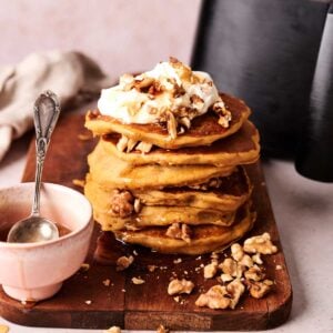 A stack of Air Fryer Pumpkin Pancake topped with whipped cream and chopped walnuts sits on a wooden board beside a small pink bowl with a spoon.