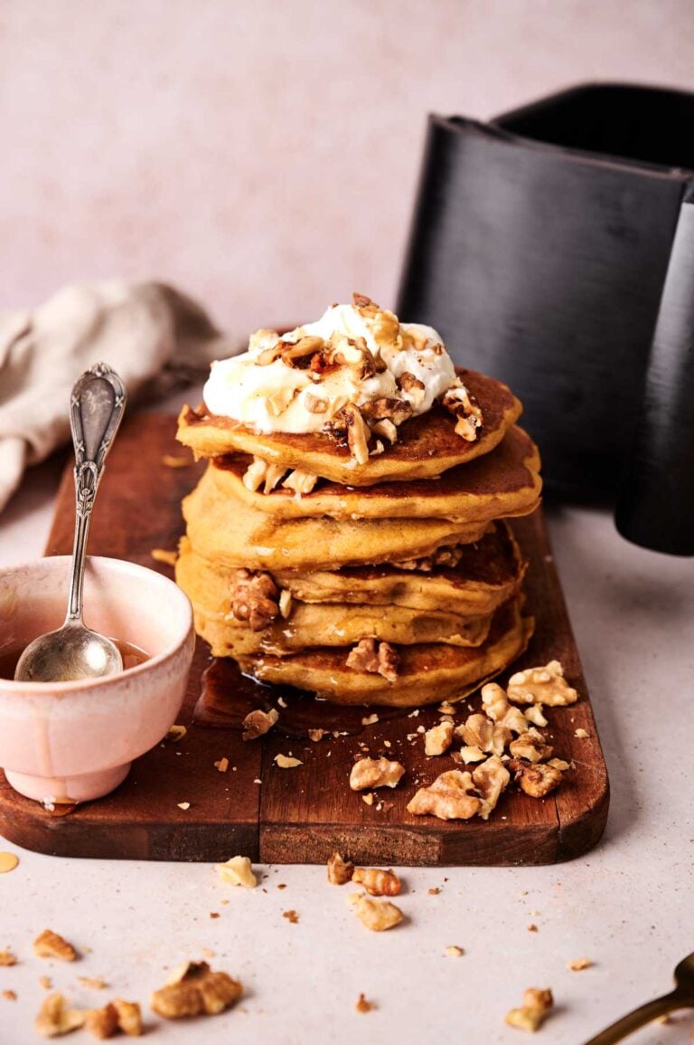 A stack of Air Fryer Pumpkin Pancake topped with whipped cream and chopped walnuts sits on a wooden board beside a small pink bowl with a spoon.