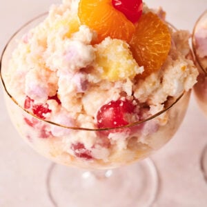 A glass dessert dish filled with classic Ambrosia Salad, made with whipped cream, marshmallows, pineapple, cherries, and mandarin oranges.