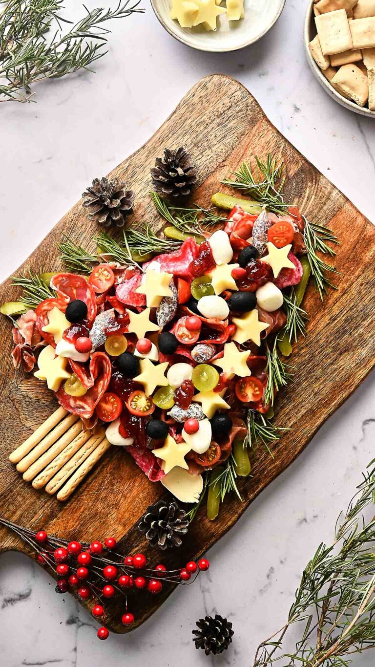A heart-shaped Christmas Tree Charcuterie Board features meats, cheeses cut into stars, tomatoes, grapes, olives, and breadsticks, all garnished with rosemary and pinecones on a wooden board.
