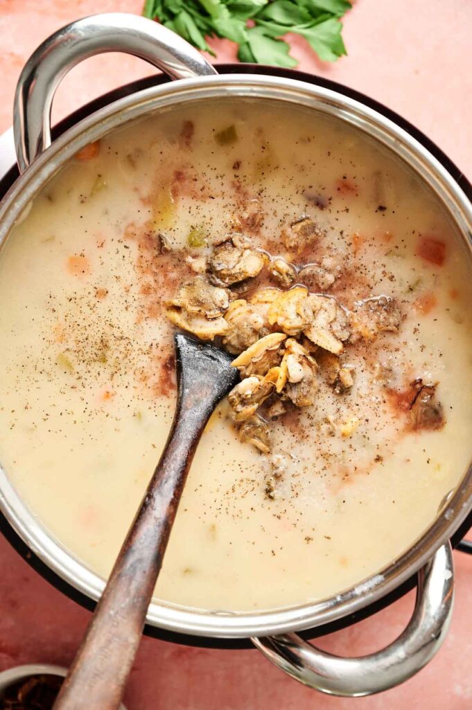 A pot of creamy clam chowder with chunks of vegetables and mushrooms, stirred with a wooden spoon and garnished with ground pepper.