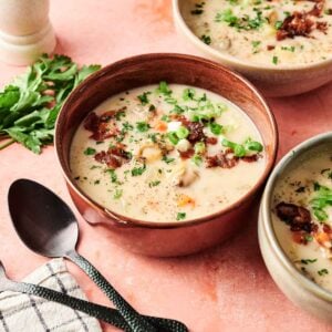 Three bowls of creamy Clam Chowder, topped with chopped green onions and crispy bacon, sit on a pink surface, accompanied by a bunch of parsley, a pepper grinder, and utensils.