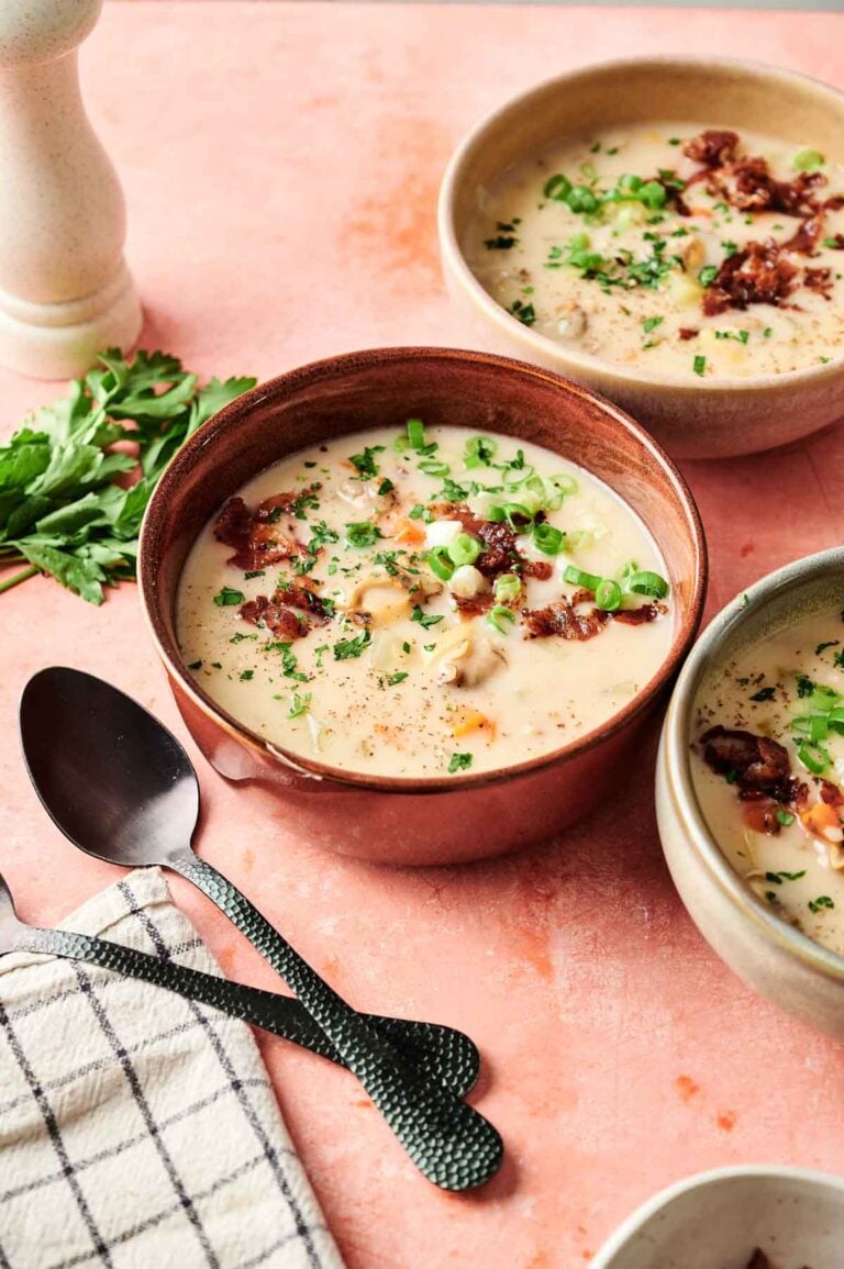 Three bowls of creamy Clam Chowder, topped with chopped green onions and crispy bacon, sit on a pink surface, accompanied by a bunch of parsley, a pepper grinder, and utensils.