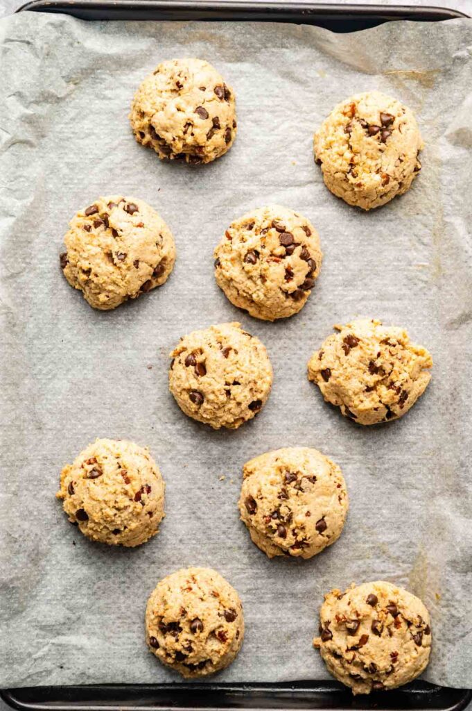 Ten scoops of unbaked Cowboy Cookies chocolate chip cookie dough are arranged on a parchment-lined baking sheet.